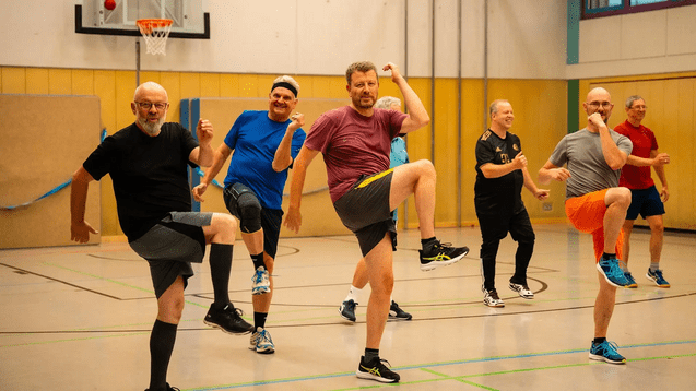 Gruppe von M&auml;nnern in Sportkleidung, die in einer Turnhalle synchron &Uuml;bungen ausf&uuml;hren.