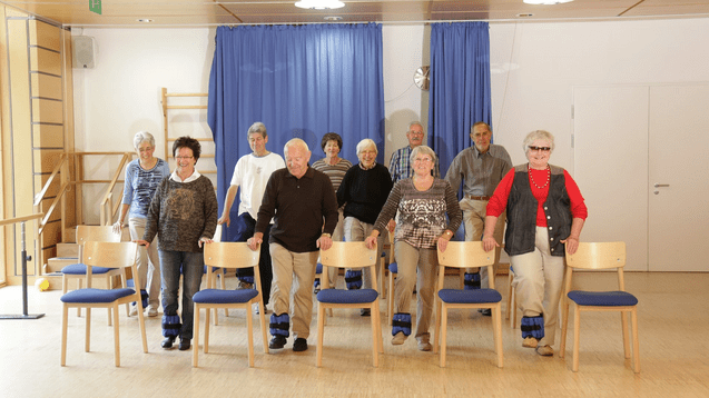 Gruppe &auml;lterer Personen steht vor St&uuml;hlen in einem &Uuml;bungsraum, einige tragen Gewichte an den Beinen.