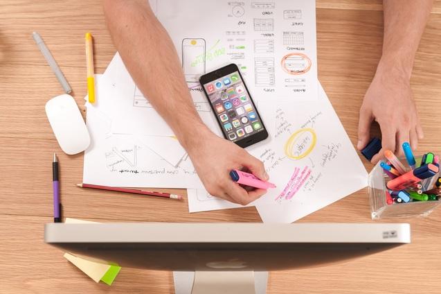 H&auml;nde mit Stiften und einem Smartphone auf einem Tisch, umgeben von Notizen und einem Computer.