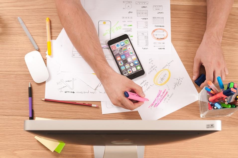 H&auml;nde mit Stiften und einem Smartphone auf einem Tisch, umgeben von Notizen und einem Computer.