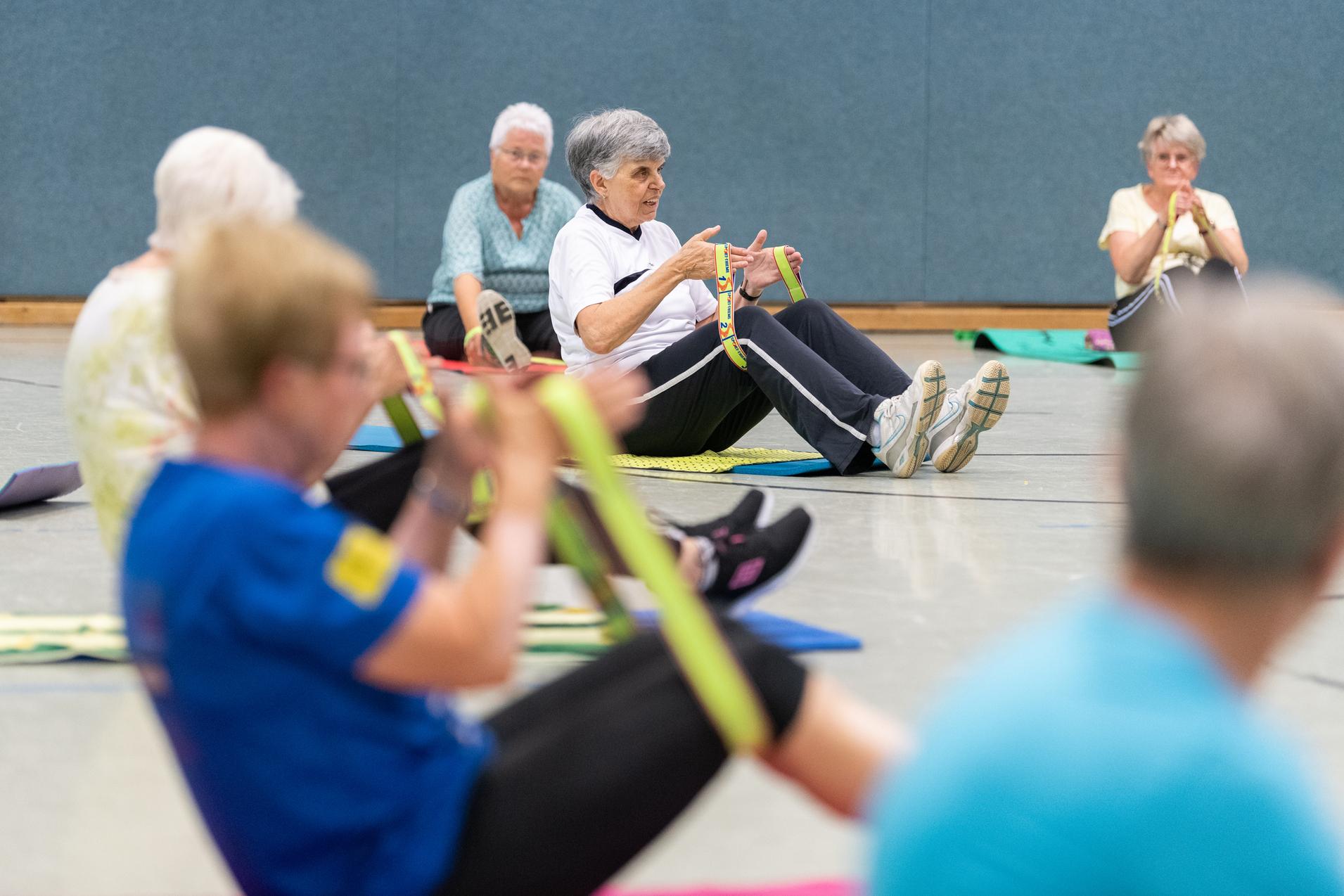Senioren nehmen an einer Fitnessübung mit Fitnessbändern in einer Sporthalle teil, sitzend auf Fitnessmatten.