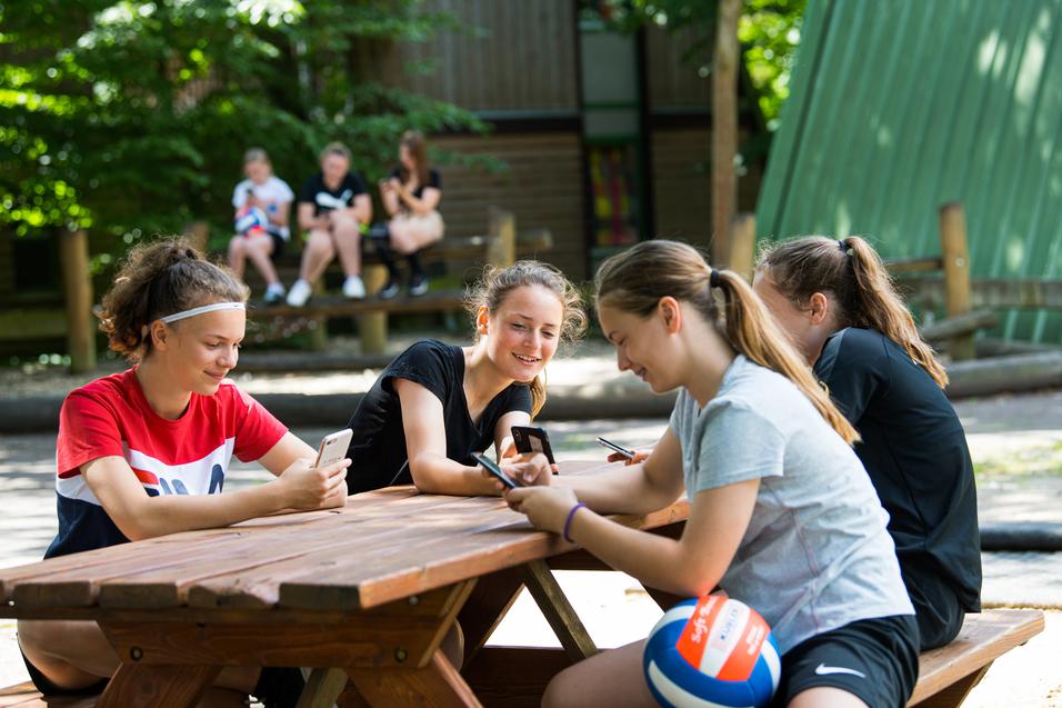 Vier Mädchen sitzen an einem Holztisch und nutzen ihre Smartphones, während im Hintergrund andere Kinder spielen.